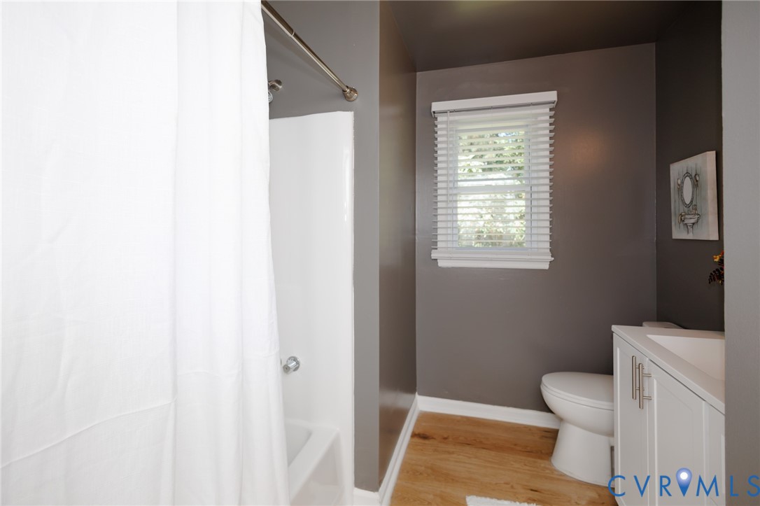 White bathtub with shower, wood-finish flooring, white vanity with integrated sink, toilet, and a window with blinds