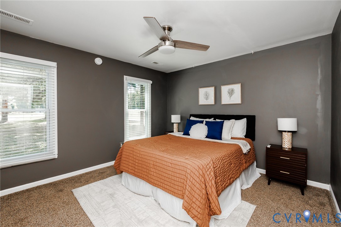 Carpeted flooring, two windows with white blinds, a ceiling fan, and painted walls