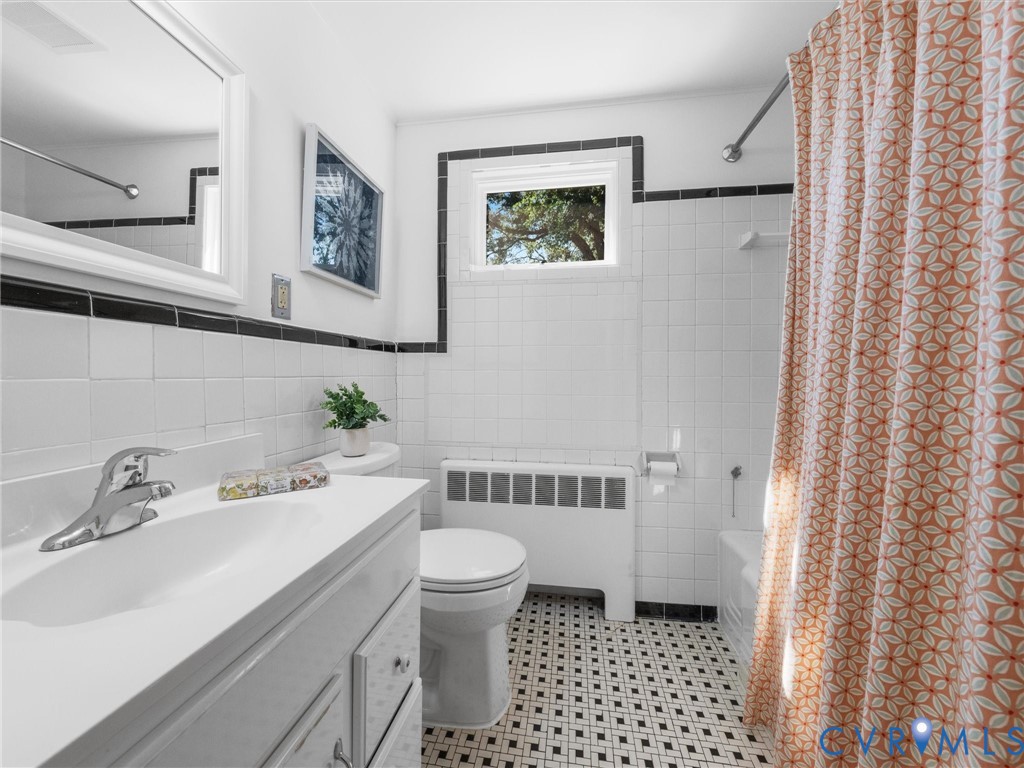 White tile walls with black accent banding, black and white mosaic tile flooring, built-in bathtub with shower, vanity with integrated sink and chrome faucet, and a window with natural light
