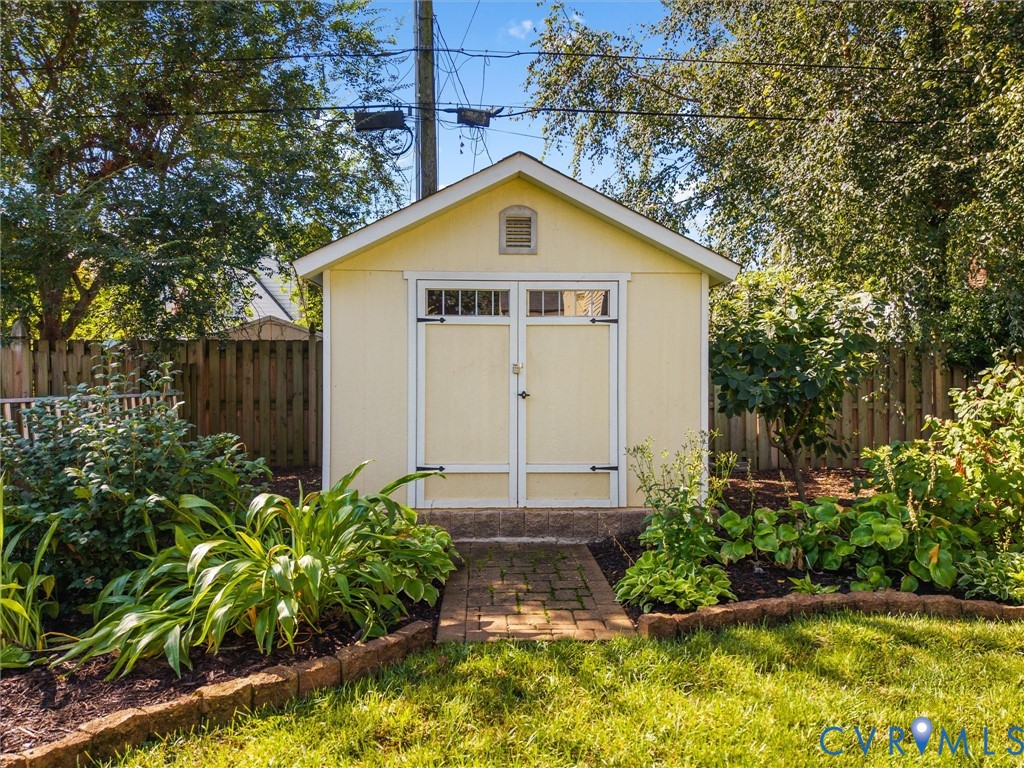Gabled storage shed featuring double doors with window panels, a brick paver pathway, and surrounding garden beds