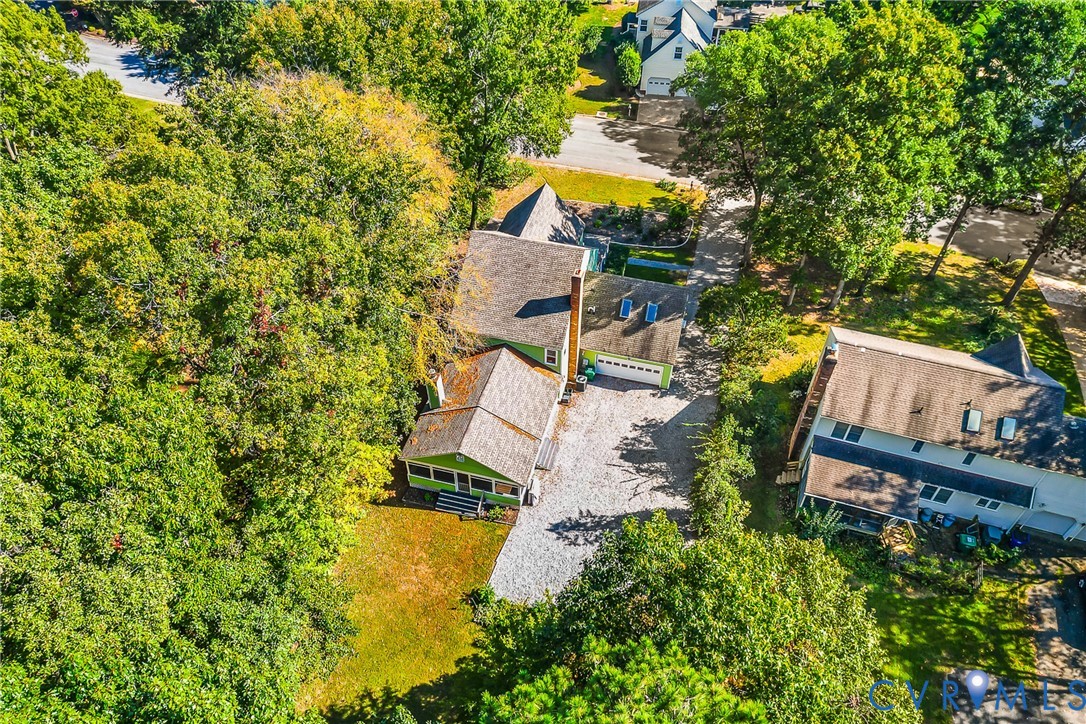 Aerial perspective featuring a multi-gabled residence with a shingled roof, attached garage, screened porch, and a stone-surfaced driveway surrounded by mature wooded landscaping