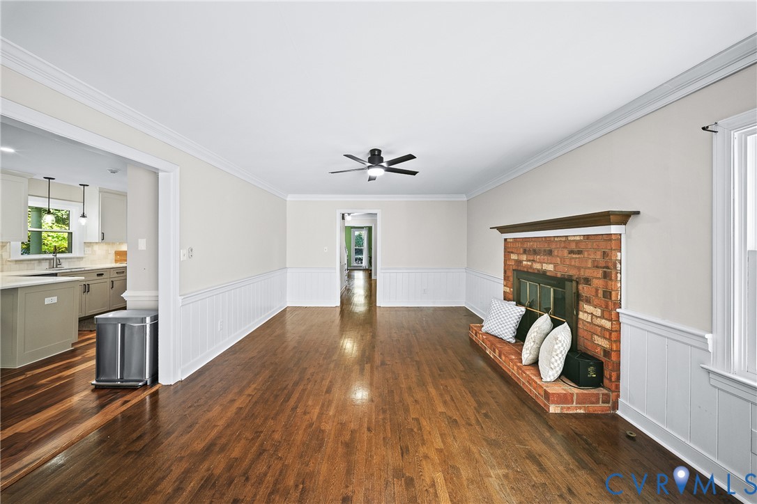 Living area featuring wood-finish flooring, brick fireplace with a wood mantel, white wainscoting, and a ceiling-mounted light fixture with fan