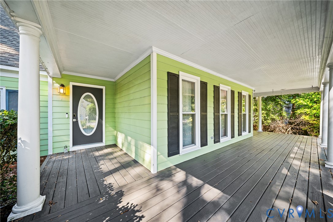 Covered front porch featuring wood-plank flooring, decorative columns, lap siding, shutter-framed windows, and an oval-insert front door Covered front porch featuring wood-plank flooring, decorative columns, lap siding, shutter-framed windows, and an oval-insert front door