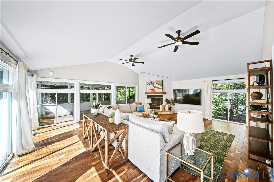 Vaulted ceilings, wood-finish flooring, two dual-blade ceiling fans, fireplace with wooden mantel, and multiple sliding glass doors