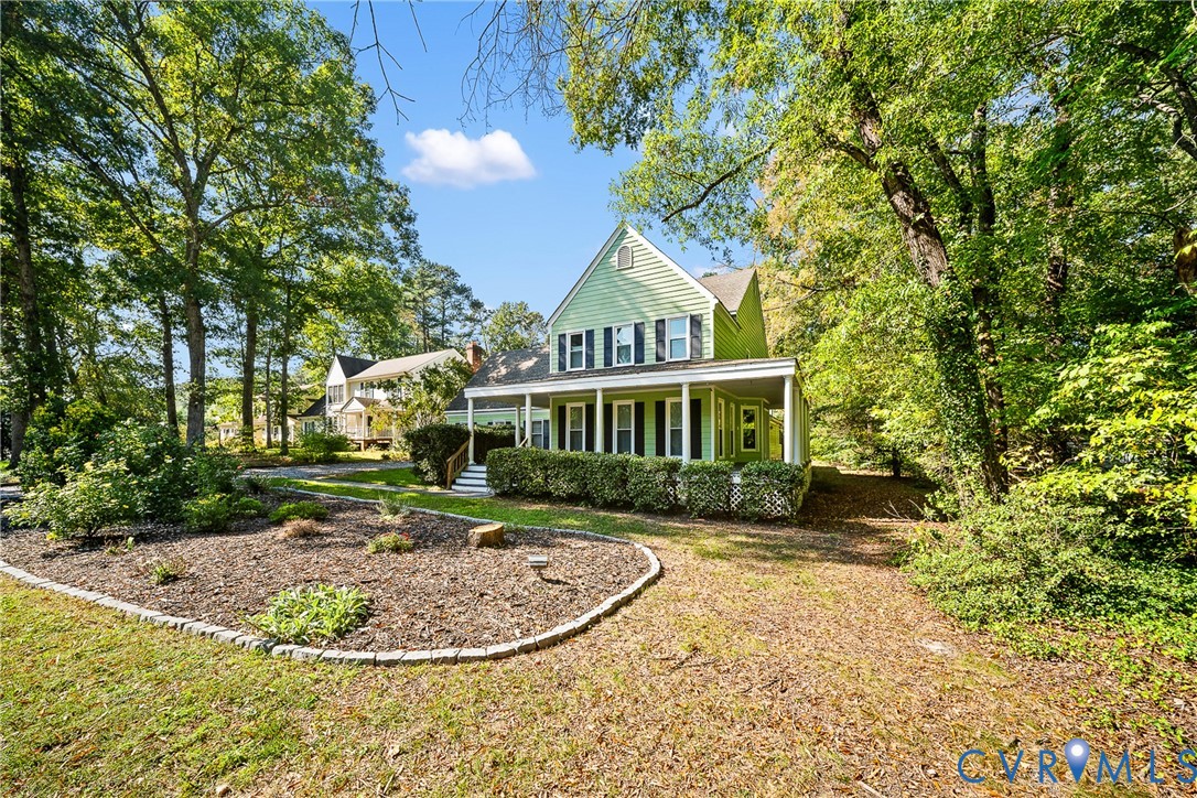 Two-story exterior featuring light green horizontal siding, dark shutters, a covered wrap-around porch, and stone-bordered garden beds situated among mature trees Two-story exterior featuring light green horizontal siding, dark shutters, a covered wrap-around porch, and stone-bordered garden beds situated among mature trees