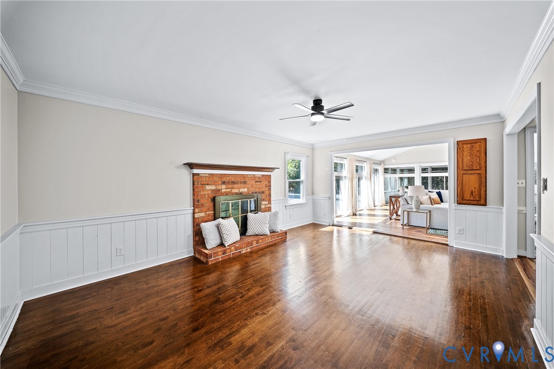 Hardwood flooring, brick fireplace with a wood mantel, white wainscoting, and a ceiling fan