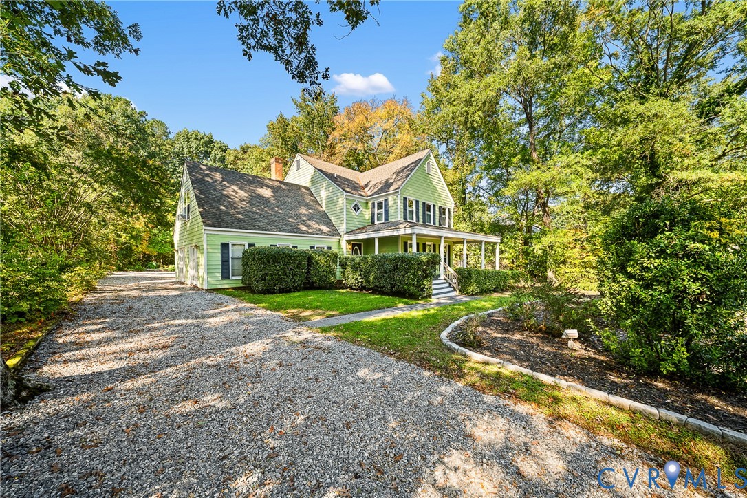 Two-story residence featuring light green siding, dark shutters, an expansive wraparound front porch, and a stone-lined garden bed Two-story residence featuring light green siding, dark shutters, an expansive wraparound front porch, and a stone-lined garden bed