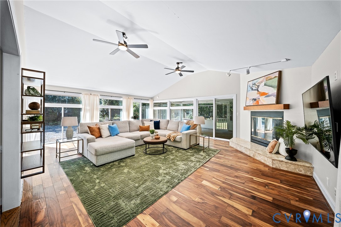 Vaulted ceilings with dual ceiling fans, expansive floor-to-ceiling windows, wood-finish flooring, and a stone-hearth fireplace with a wood mantel
