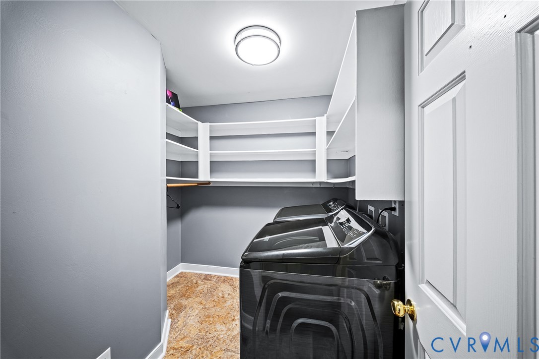 Laundry space featuring multi-tiered white built-in shelving, flush-mount ceiling lighting, textured stone-pattern flooring, and a paneled door with brass-finish hardware