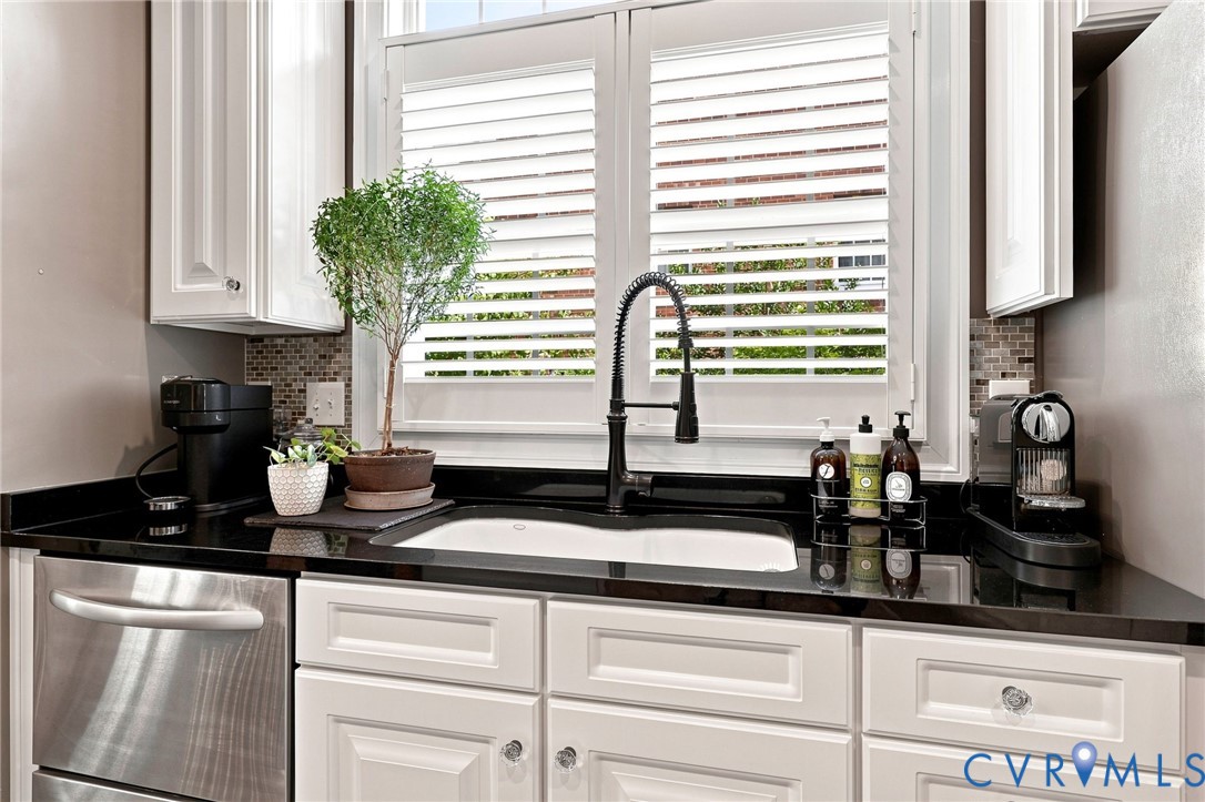 White cabinetry with crystal knobs, dark countertops, tile backsplash, stainless steel dishwasher, and a window with plantation shutters