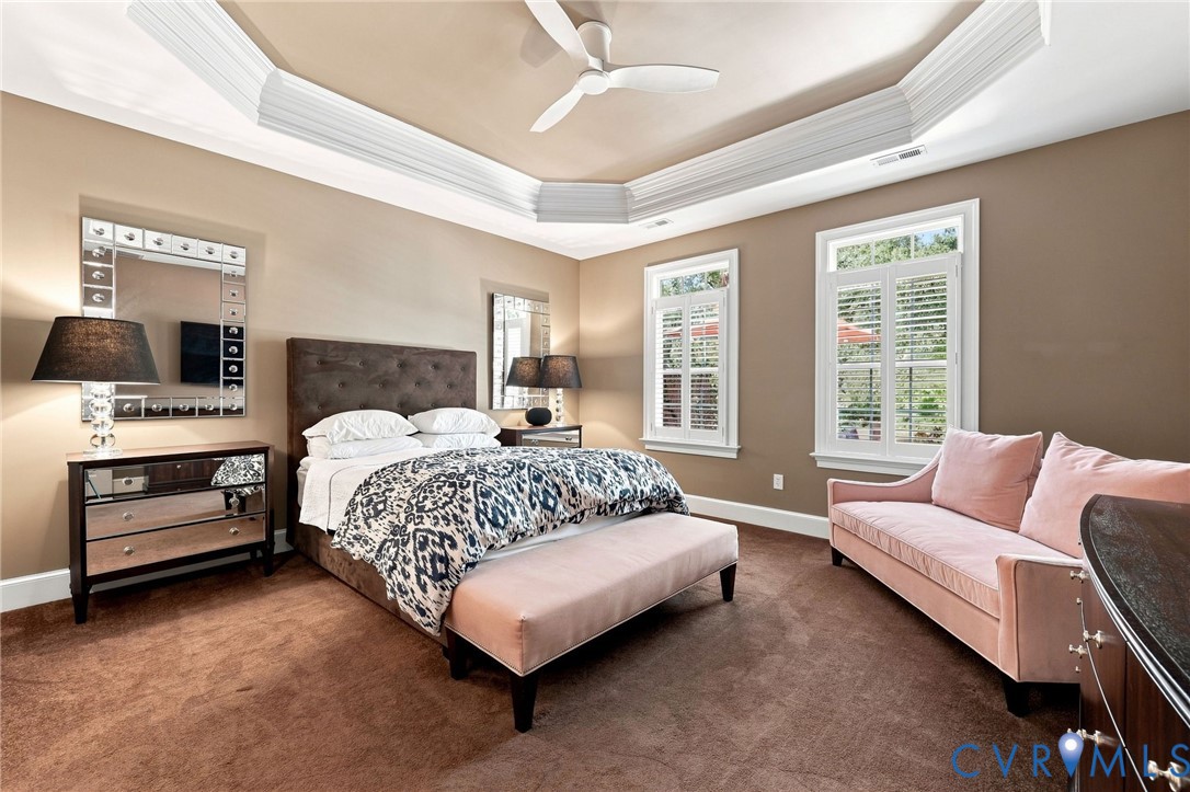 Tray ceiling with crown molding, ceiling fan, two windows with plantation shutters, mirrored dresser, and wall-to-wall carpeting