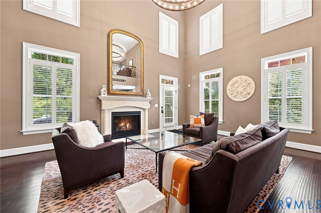 Soaring ceilings with upper-level windows, a stone-surround fireplace, dark wood-finish flooring, and white plantation shutters on multiple windows