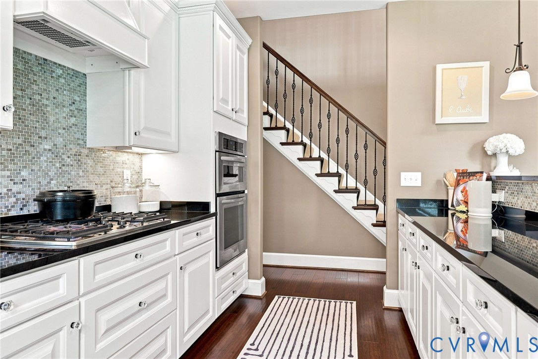 White cabinetry with polished hardware, dark countertops, a mosaic tile backsplash, built-in double ovens, and wood-finish flooring