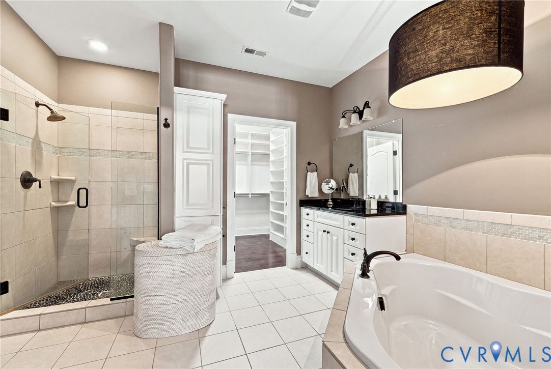 Spacious bathroom featuring a built-in soaking tub, a separate tiled shower with a glass enclosure, white vanity cabinetry with a dark countertop, and a large walk-in closet
