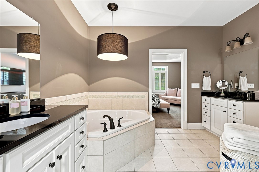 Corner soaking tub with tiled surround, dual vanity with undermount sinks, and ceramic tile flooring