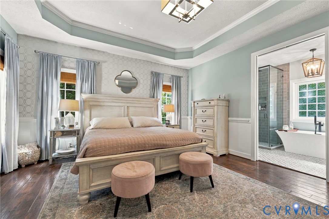 Tray ceiling with crown molding, wood-finish flooring, two large windows, white paneled wainscoting, and an en-suite bathroom with a freestanding tub and glass-enclosed shower
