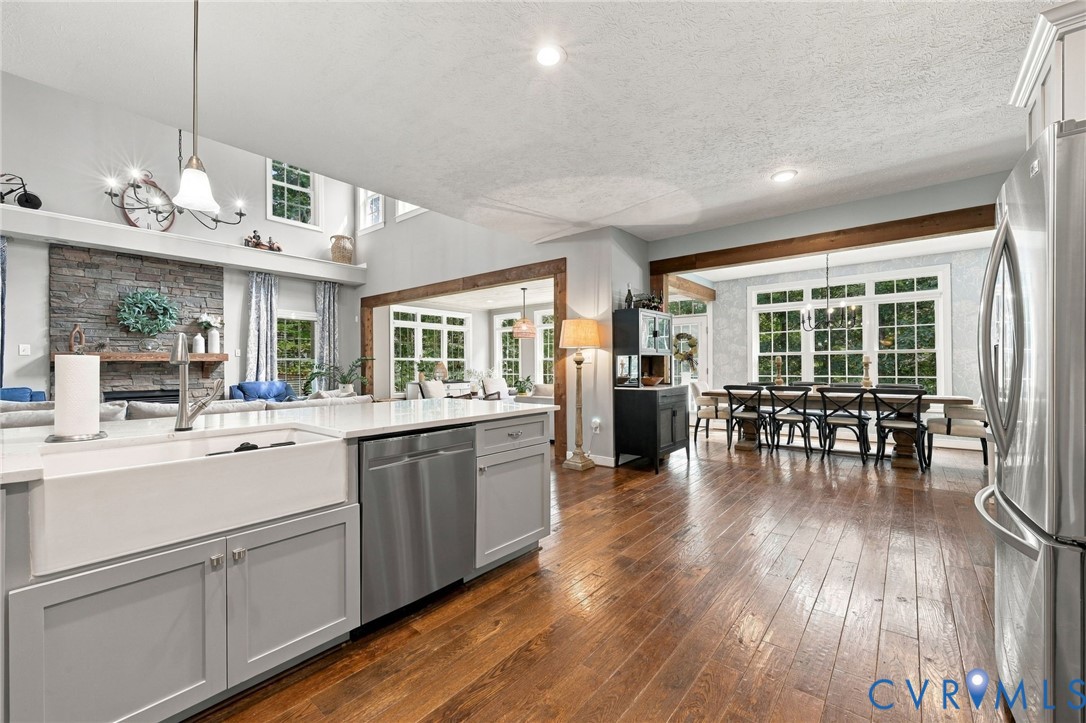 Open-concept living area featuring wood-finish flooring, a stone-faced fireplace with a mantel, a farmhouse sink, stainless steel appliances, and exposed wood beams