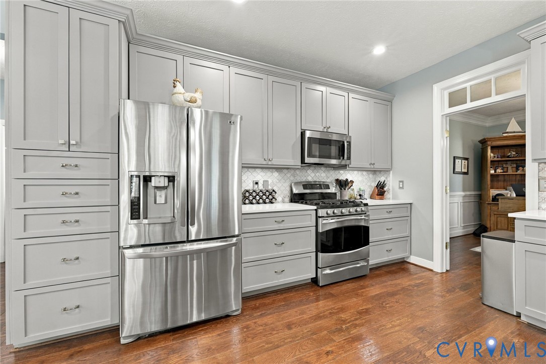 Light gray cabinetry, stainless steel appliances, mosaic tile backsplash, white countertops, and wood-finish flooring