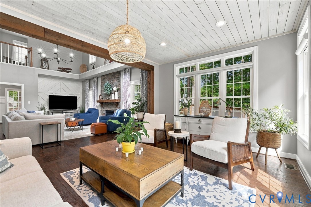 Spacious living area featuring exposed wood beams, wood-finish flooring, a stone fireplace, a white plank ceiling, and large multi-pane windows