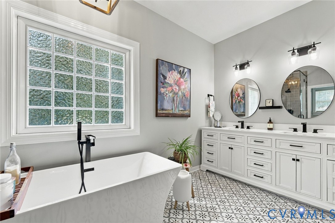 Freestanding soaking tub with a floor-mounted matte black faucet, frosted glass block window, double vanity with white cabinetry, dual undermount sinks, and patterned floor tile