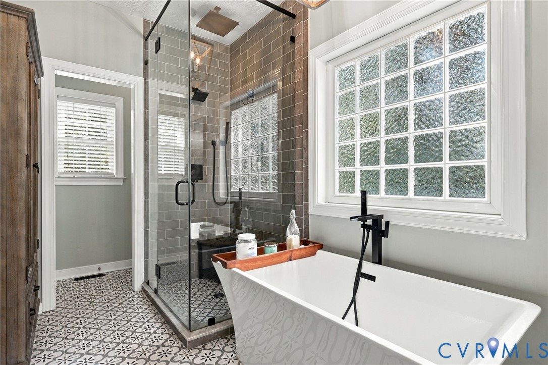 Freestanding soaking tub with a floor-mounted matte black faucet, glass block window, walk-in shower with tile surround, and patterned floor tile