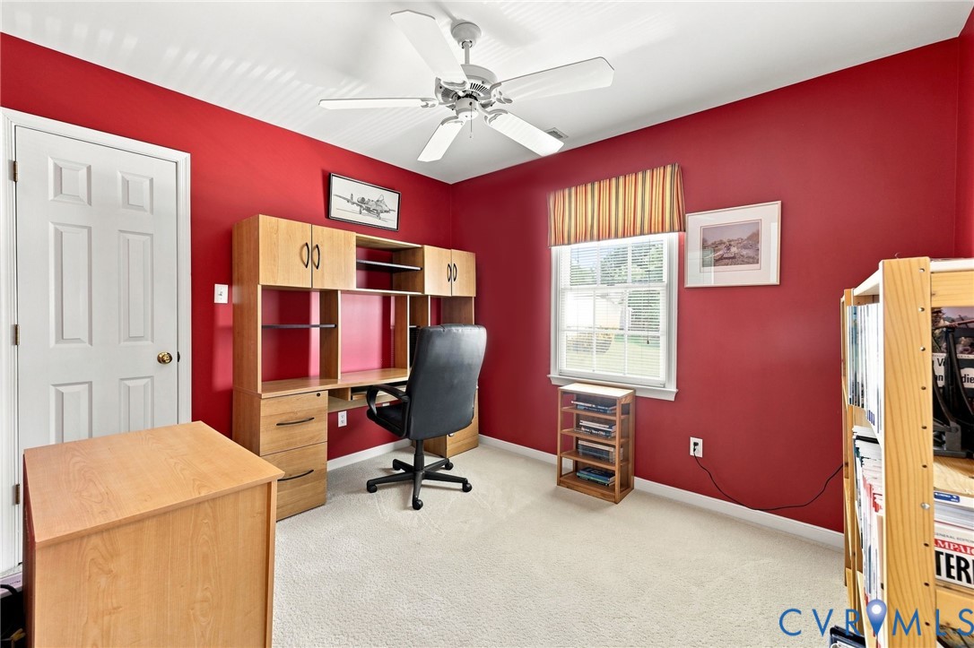 Bright, versatile bedroom featuring vibrant red walls, wood-finish cabinetry with built-in shelving and storage, carpet flooring, ceiling fan, and abundant natural light from large windows