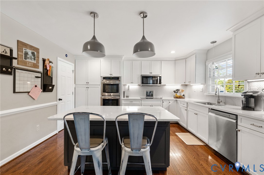 White cabinetry, stainless steel appliances, wood-finish flooring, central island with seating, and dual pendant lighting fixtures White cabinetry, stainless steel appliances, wood-finish flooring, central island with seating, and dual pendant lighting fixtures