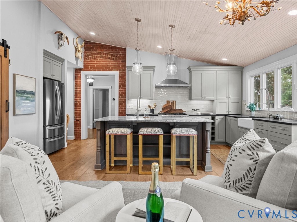 Kitchen featuring an angled wood-paneled ceiling, stainless steel appliances, a center island with pendant lighting, a farmhouse sink, and a brick wall accent