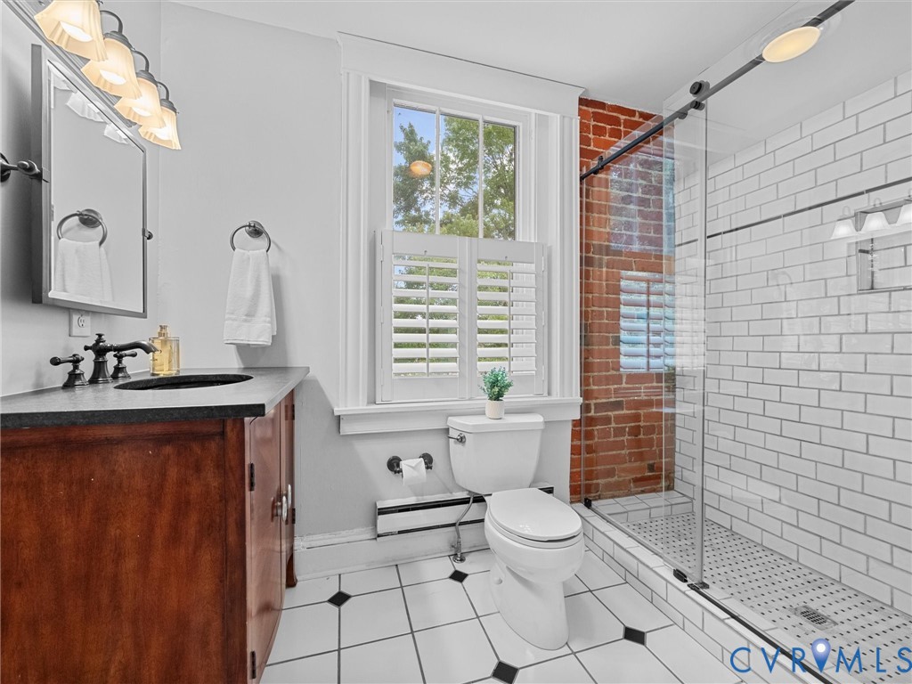 Bathroom featuring a glass-enclosed shower with white subway tile, exposed brick wall, wood vanity with dark countertop, and a window with plantation shutters