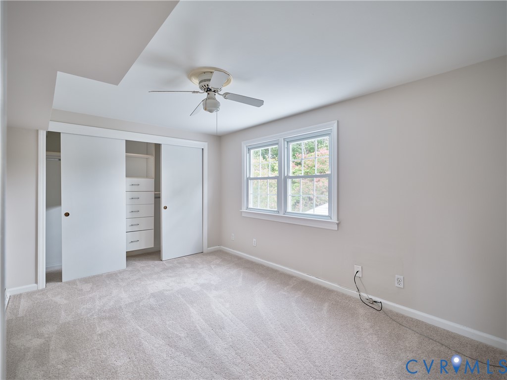Plush carpet flooring, white trim details, window providing natural light, ceiling fan fixture, and custom closet built-ins