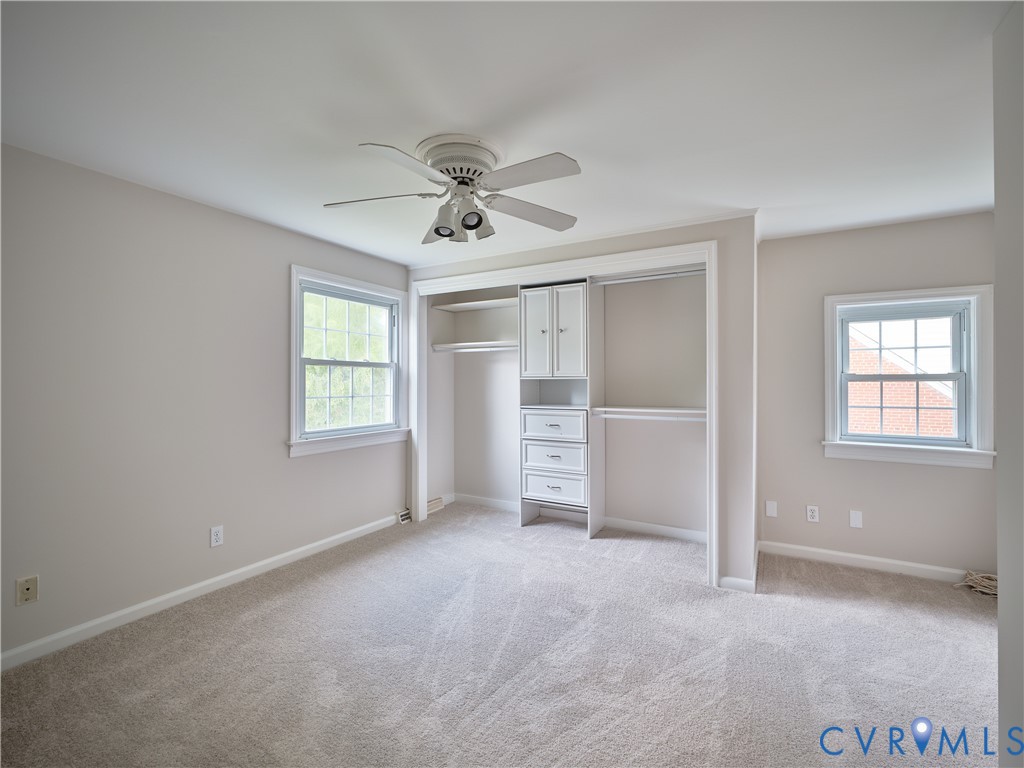 Plush carpet flooring, ceiling fan with integrated lighting, two windows with white trim, and a built-in closet system featuring drawers and cabinetry