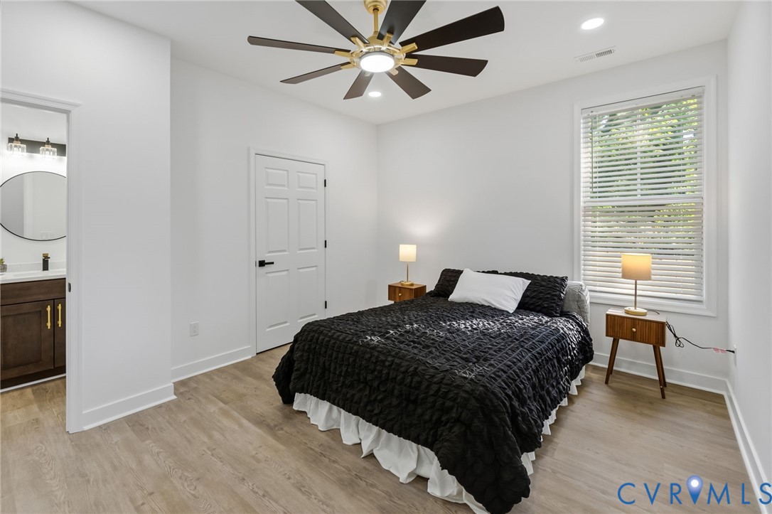 Bright secondary bedroom featuring wood-finish flooring, a contemporary ceiling fan with integrated lighting, large operable window with plantation shutters, and direct access to an ensuite bathroom