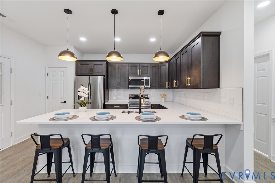Kitchen featuring dark wood-finish cabinetry, stainless steel appliances, a white subway tile backsplash, and a large island with a light-toned countertop and pendant lighting