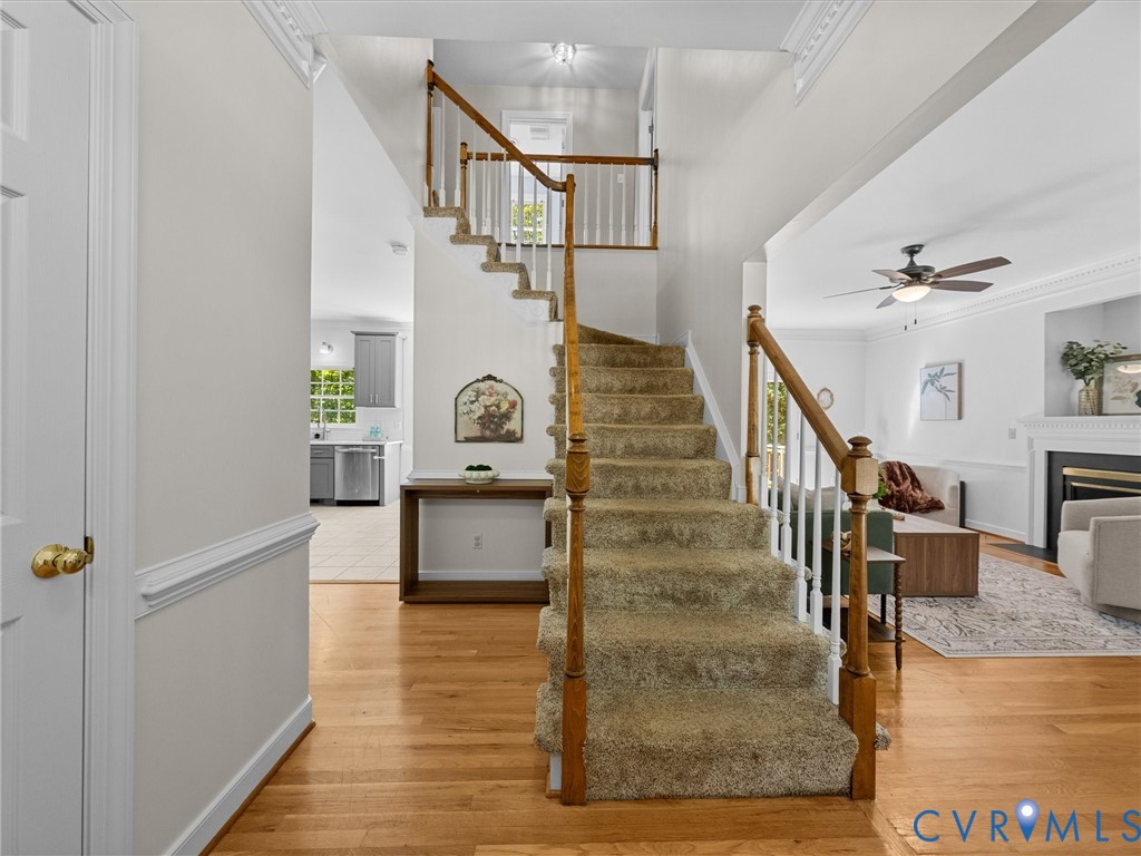 Hardwood-style flooring throughout the main level, staircase with carpeted treads and wood banister, a living area with a ceiling fan, and a fireplace with a white mantel Hardwood-style flooring throughout the main level, staircase with carpeted treads and wood banister, a living area with a ceiling fan, and a fireplace with a white mantel