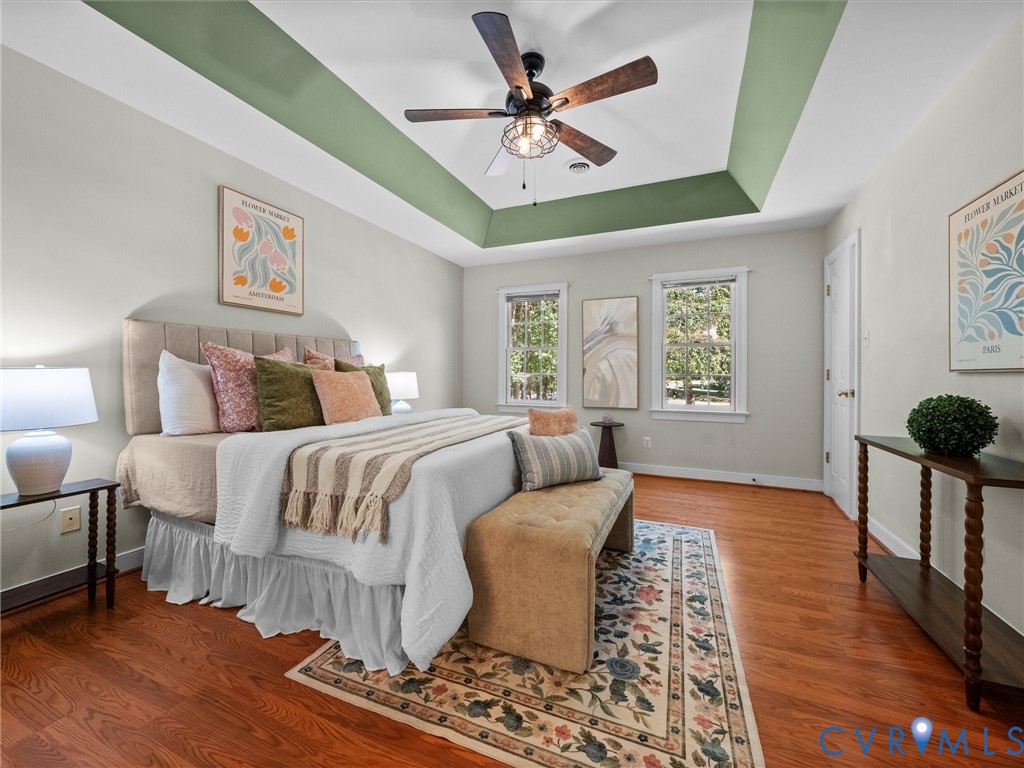 Tray ceiling with accent paint, wood-finish flooring, two windows providing natural light, ceiling fan with light fixture, and a solid white door Tray ceiling with accent paint, wood-finish flooring, two windows providing natural light, ceiling fan with light fixture, and a solid white door