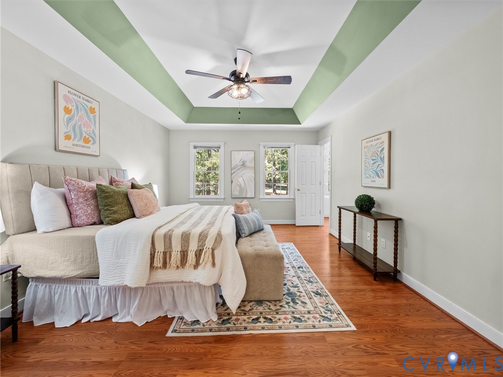 Tray ceiling with decorative paint accent, ceiling fan with integrated lighting, wood-finish flooring, white paneled interior door, and multiple windows