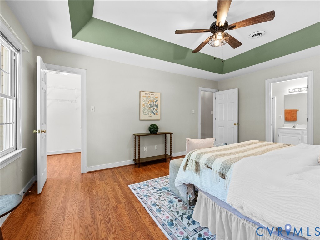 Hardwood flooring throughout, tray ceiling with a ceiling fan, white paneled doors, and an en-suite bathroom with a vanity