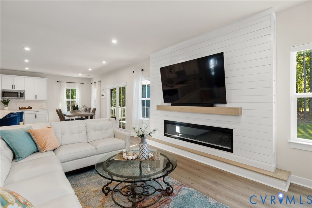 Open-concept living space featuring a linear electric fireplace with a wood mantel, white shiplap accent wall, recessed lighting, and wood-finish flooring