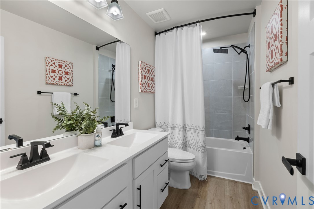 Dual vanity with matte black fixtures, full-width mirror, light wood-finish flooring, tiled bath and shower combination, and built-in toilet