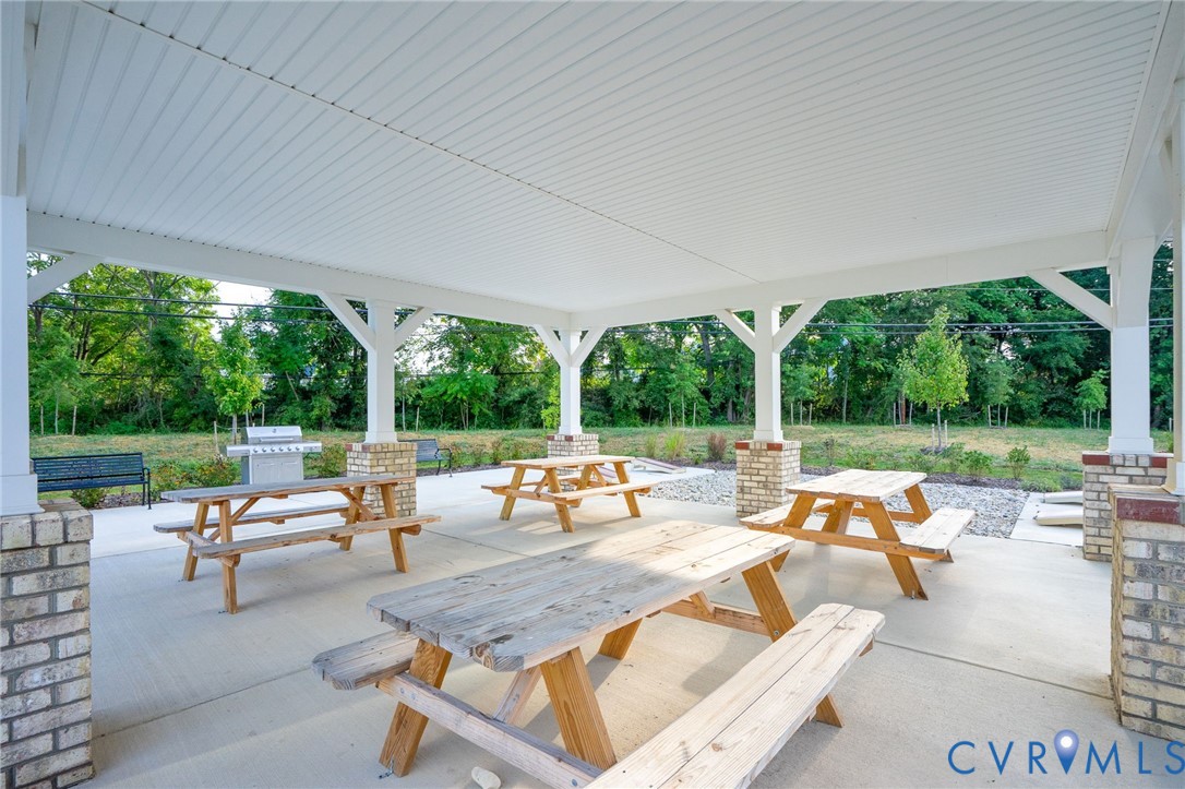 Covered outdoor patio with a concrete surface, supported by brick-clad pillars, and featuring multiple picnic tables and a stainless steel grill