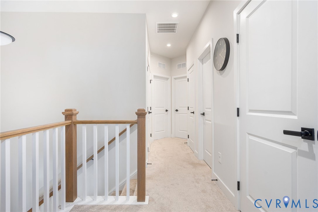 Light-colored carpet flooring, white paneled doors with matte black hardware, recessed lighting, and a light-toned wooden banister with white balusters