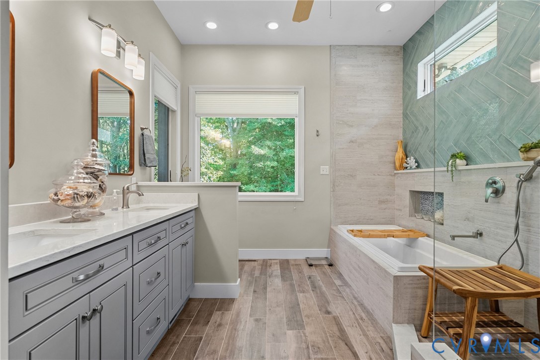 Dual vanity with gray cabinetry and white countertops, wood-finish flooring, built-in soaking tub, glass-enclosed shower with tiled walls, and recessed lighting
