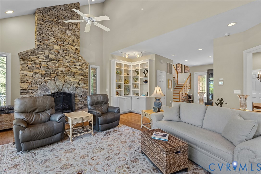 Vaulted ceiling with a ceiling fan, stone fireplace, built-in shelving, hardwood flooring, and a staircase with a wood handrail
