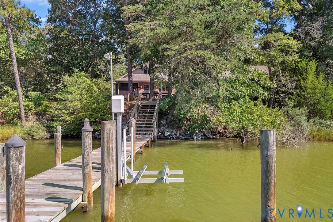 Private wooden dock featuring a boat lift, leading to a waterside property with mature trees and a visible residential structure