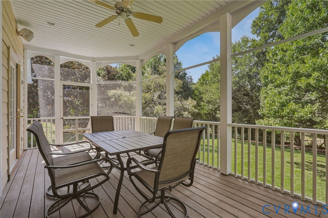 Expansive screened porch with a wood-finish deck, ceiling fan, and white railing overlooking a grassy area with mature trees