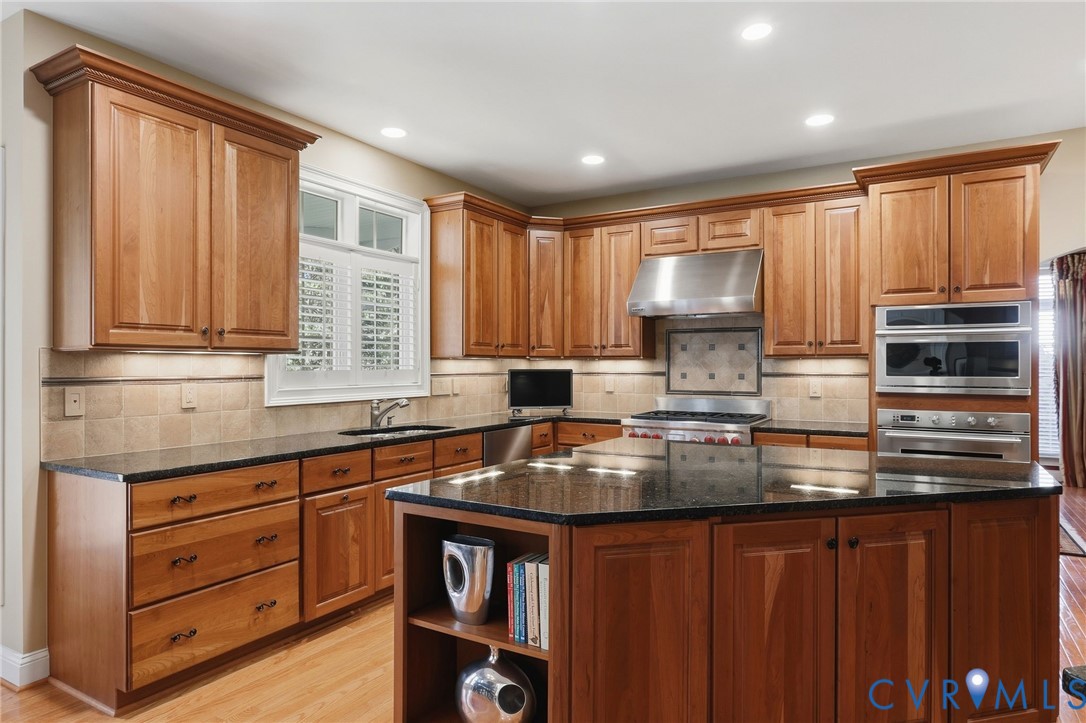 Kitchen featuring wood cabinetry, dark countertops, a large island with shelving, stainless steel appliances, and a tiled backsplash Kitchen featuring wood cabinetry, dark countertops, a large island with shelving, stainless steel appliances, and a tiled backsplash