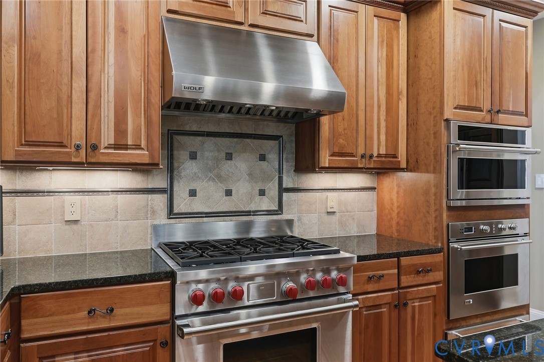 Stainless steel range with a professional-grade hood, built-in wall oven and microwave, stone countertops, and wood cabinetry with under-cabinet lighting