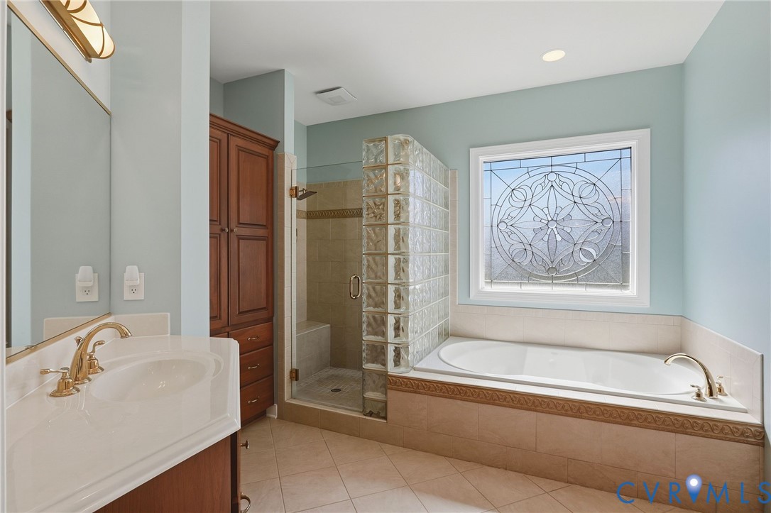 Soaking tub with decorative tile surround, walk-in shower with glass block accent, custom stained-wood cabinetry, and an ornate leaded-glass window