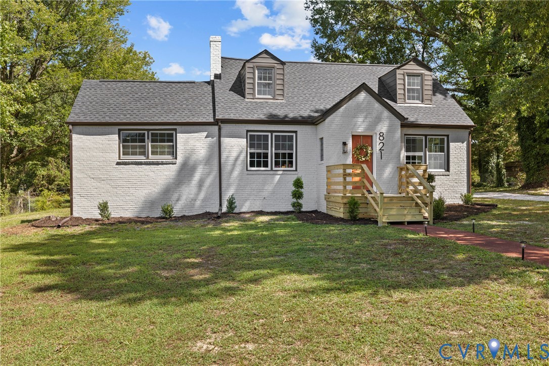 White brick exterior with dark trim windows, front porch with wooden railings, dark shingled roof, and dormer windows
