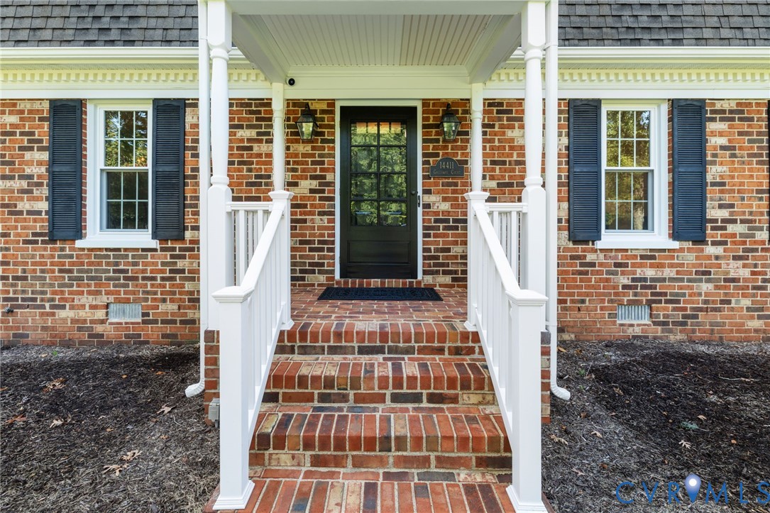 Brick exterior featuring a covered porch with white columns, a dark panel door with glass inserts, and white window frames with dark shutters